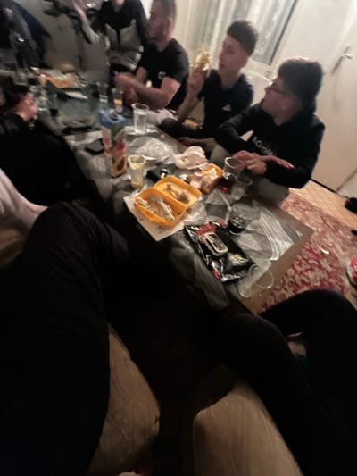 A low-angle, close-up photograph captures a group of people gathered around a low table filled with food and drinks. The setting appears to be indoors, possibly in a residential space given the presence of a rug and sofa. Three young men are visible on the left side of the table, they are eating. The table is covered with various food items, including what appears to be tacos, snacks, and drinks. Several clear plastic cups are scattered across the table. A person is sitting on a light-colored sofa and wearing black pants. The lighting in the room is dim, casting shadows and affecting the clarity of details. The image does not seem to be located in Lenovo, Bulgaria.