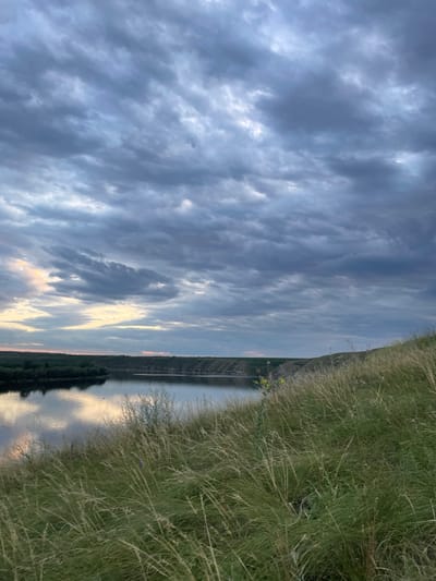 A landscape image shows a body of water, likely a river or lake, under a cloudy sky. The sky occupies most of the frame with layered, textured clouds in shades of gray and blue. Along the right edge, tall grass fills the foreground, slightly blurred. The water reflects the sky's colors and the distant treeline on the opposite shore. Overall, the scene evokes a tranquil, slightly overcast evening.