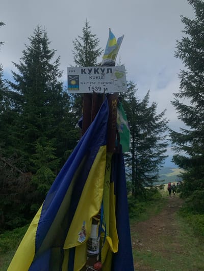 This image depicts the summit marker for Mount Kukol (Kukyl), a peak in the Carpathian Mountains near Vorokhta, Ukraine, identified by the coordinates N 48°12'56", E 24°32'43" and an elevation of 1539 meters.

The central subject is a sturdy, dark-colored pole, likely wood or metal, with a white rectangular sign affixed at its top. The sign displays "КУКУЛ" (KUKUL) in Cyrillic, "KUKUL" in Latin script, the geographic coordinates, and the elevation "1539 M". A smaller yellow and blue sign to the left features the text "НЕ СТІЙ НА ШЛЯХУ" (Do Not Stand on the Way) and a stylized symbol.

Multiple flags are draped around and attached to the pole. A large blue and yellow flag, representative of Ukraine, is prominently wrapped around the lower portion of the pole, while another, smaller Ukrainian flag is visible at the very top. A green and white flag, along with other less distinct banners, are also affixed. Below the main flags, various small objects, including a cylindrical item wrapped with string and some red components, are tied to the pole.

The environment is a mountainous landscape. Tall, dark green coniferous trees frame the scene on both sides and in the background, suggesting a dense forest at elevation. The sky is overcast with light gray clouds. A dirt trail or path extends into the distance on the right side of the image. Along this path, two to three individuals, appearing to be hikers with backpacks, are walking away from the camera into the receding perspective of the trail and the valley beyond. The ground immediately surrounding the pole is covered in green grass and low vegetation.