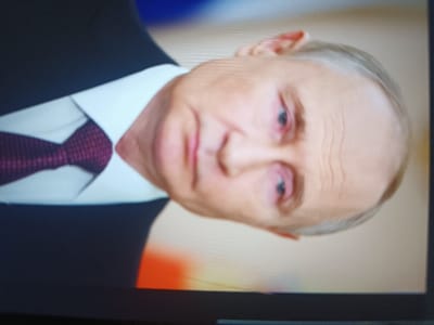 The image shows a close-up of Vladimir Putin. He is wearing a black suit, a white shirt, and a dark red patterned tie. His gaze is directed toward the viewer. His skin shows signs of aging with wrinkles. His eyes appear tired, possibly bloodshot. The background is blurred. The image appears to be a photograph taken of a screen.