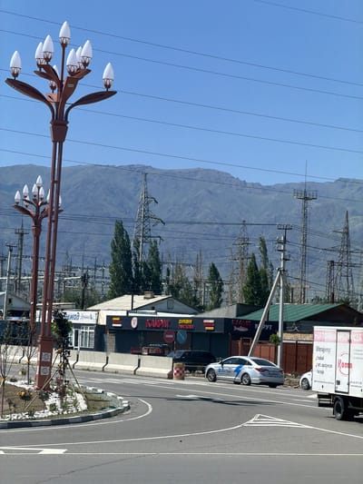 The image captures an outdoor scene, likely in Balykchy, Kyrgyzstan, on a clear day with bright sunshine. The foreground features a decorative lamppost with multiple white, oval-shaped lights. In the middle ground, a white taxi with blue accents and the text "Navi Taxi" is parked on the side of a paved road. Adjacent to the road are several small buildings with visible signage in Cyrillic script, including "KAUPAT" and what appears to be a fish market or shop ("БАЛЫК КРУТ"). A white delivery truck with "HURTOWNIA" and "Roma" written on its side is also visible. In the background, a vast mountain range dominates the horizon, and numerous tall electrical pylons and power lines crisscross the landscape, suggesting an area with significant infrastructure. The scene appears to be a commercial or roadside area, possibly near a transportation hub, given the presence of the taxi and delivery truck. The overall impression is one of a functional, everyday setting under a clear blue sky.