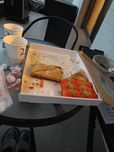 The image captures a close-up view of a casual outdoor setting on what appears to be a balcony or patio, featuring partially eaten desserts and beverages on a glass-top table.

In the center of the frame, a white cardboard box holds various Middle Eastern pastries. On the left side of the box, there's a large, golden-brown, flaky pastry, possibly a type of baklava, which appears to be partially eaten, revealing some orange-colored filling or residue underneath. To the right, a vibrant orange, rectangular pastry, likely kunefe (knafeh), is visible, generously topped with crushed green pistachios. A smaller, triangular piece of flaky pastry, also adorned with pistachios, is situated behind the kunefe. The white box liner shows scattered green pistachio crumbs and orange remnants from the pastries.

To the left of the pastry box, two white paper cups are placed on the table. The front cup clearly displays a logo with "2 cups" and "Dantes," along with text indicating "Columbian Coffee," "Tea," and other partially visible words like "Arizon" and "Club." A small, dark-colored tea bag or similar item rests on the table next to this cup. Beside the cups, a crumpled white paper bag or napkin is visible, along with several crumpled tissues or napkins that have red stains, possibly from the pastries or another food item.

The table itself is a glass-top design with a dark metal frame. In the background, a dark, woven outdoor chair is partially visible behind the table. The floor appears to be tiled, consistent with an outdoor space. To the far left in the background, a dark, wheeled object, possibly a small utility cart, can be seen. On the right side of the image, beyond the table, there's a light-colored wall or building exterior. A tall window or sliding glass door reflects a warm, golden light, suggesting it might be late afternoon or early evening.

On the right edge of the table, a brown cardboard box, possibly the carrier for the pastry box, is visible, with some text that appears to be in Hebrew script (including "מתוקים" which means "sweets"). A white woven basket is placed on this brown box.

At the very bottom of the image, two feet wearing dark, moccasin-style slippers with fur lining are visible, indicating that someone is seated at the table, enjoying the treats and drinks. The scene suggests a relaxed moment of indulgence outdoors.