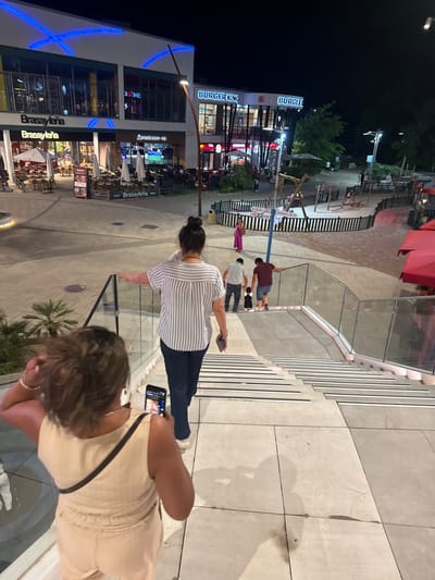 The image shows an outdoor urban scene at night, likely a pedestrianized entertainment or shopping area. A Burger King restaurant is visible with its signature signage. Several people are walking down a wide, modern outdoor staircase, which is illuminated. In the foreground, a person is holding a smartphone and appears to be taking a photo. Another person, seen from behind, is also on the staircase, wearing a striped shirt and dark pants. Further down the stairs, a family, including a man, a woman, and a small child, is descending. To the left of the stairs, outdoor seating areas with umbrellas are set up in front of businesses labeled "Brasayleña." The overall environment suggests a vibrant evening atmosphere in Alcorcón, Spain, likely in a commercial district with dining and leisure options.