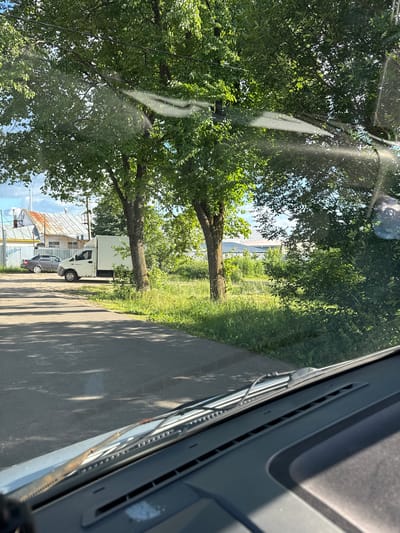 The image shows a view from inside a vehicle, looking out at a street scene in Yuzha, Russia. The foreground is dominated by the car's dashboard and windshield, with windshield wipers visible. Through the windshield, a paved road stretches into the distance, bordered by green grass and trees.
On the left side of the road, there is a silver sedan parked. Next to it is a white cargo van. Behind the vehicles, a fence runs along the property line, and beyond that, buildings with white walls and metal roofs are visible. The sky is partly cloudy, with blue sky peeking through. The overall impression is of a quiet, residential or industrial area on a sunny day. The trees are lush and green, suggesting a warm season. There are no people visible in the image. The lighting indicates it is daytime, likely afternoon, given the angle of the sun and the shadows cast by the trees.