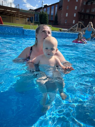 A woman and a toddler are in a swimming pool, with the woman holding the toddler. The toddler is facing the camera and has blue eyes. The woman is looking to the side. Other people are visible in the pool in the background. The pool is blue and clear, with sunlight reflecting off the surface. Buildings and trees are visible in the background. The location appears to be an outdoor swimming pool complex.