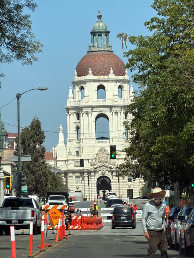In Pasadena, California, the Pasadena City Hall is visible in the background, a large, ornate building with a prominent dome and arched windows. In the foreground, a man wearing a cowboy hat and a light-colored jacket walks across a street. Construction is underway, with orange traffic barriers, cones, and a "LANE CLOSED" sign visible. Several vehicles, including a Ford pickup truck, are present in the scene, some of which appear to be stopped due to the road work. Traffic lights are visible, with one showing a green signal. Trees line the street, with some branches extending into the frame. The sky is clear and blue.