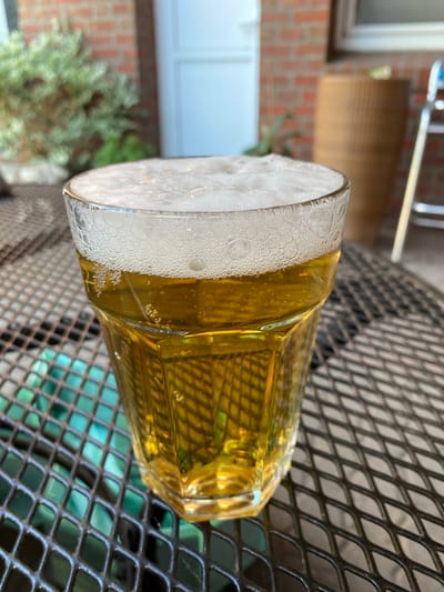 A glass of beer is resting on a dark, diamond-patterned metal table. The beer is a golden color and topped with a thick layer of white foam, featuring many small bubbles. The glass itself is clear and appears to be made of thick glass, with angular facets. Sunlight is glinting off the surface of the beer, suggesting it is daytime.
In the background, out of focus, is a brick wall with some greenery and a white door or window. A metal railing or structure is visible above the brick wall. The overall scene appears to be outdoors, possibly on a patio or balcony, given the table and the background elements. There is no discernible text in the image. The lighting and the presence of a beverage suggest a relaxed, possibly social, moment during the day. The location context of Lütjenwestedt, Germany, doesn't provide specific visual cues within the image itself but places the scene geographically.