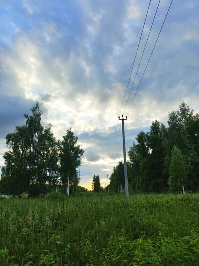 This media file shows a rural landscape in Tsibino, Russia, at what appears to be either sunrise or sunset, given the golden light on the horizon. The scene is dominated by nature, with lush green grass and trees filling the right and center portions of the frame. The sky is a mixture of blue and white clouds, with streaks of sunlight breaking through, suggesting dynamic weather.
In the foreground and midground, tall grass and various plants are visible, indicating a natural, uncultivated area. Along the left side of the image, a line of trees, predominantly birch trees with their distinctive white bark, forms a border. A single power line pole with insulators and wires stretches vertically through the scene, a man-made element contrasting with the natural surroundings. The wires are taut and extend across the sky. Further in the distance, another, smaller power line pole is visible.
The overall mood is tranquil and serene, with the natural elements conveying a sense of peace. The lighting suggests a transitionary time of day, adding a dramatic or contemplative quality to the image. There is no visible text in the media file.