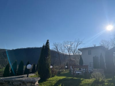 This image captures a serene outdoor scene in Pishtane, Bulgaria, under a clear, bright blue sky. The sun, prominently featured in the upper right, radiates intense light, creating noticeable lens flare that stretches across the upper half of the frame.

The setting is a residential garden with a house in the midground, backed by undulating hills or mountains. The distant hills are densely covered with dark trees, appearing a deep green or shadowed hue. In front of these hills, patches of lighter green fields are visible.

The house itself is a light-colored, possibly off-white, two-story structure with a dark, sloped roof and a visible window on its side. A dark door or entryway is also visible. The surrounding garden features several tall, slender evergreen trees, likely cypresses, lining paths and standing near the house. Interspersed among these are bare deciduous trees, suggesting a season of late winter or early spring.

In the immediate vicinity of the house, a wooden outdoor table is set with white chairs on a patch of green grass. Spherical white garden lights on poles are dotted throughout the garden, with one particularly prominent in the left foreground, atop a low stone wall. The grass is lush and green, marked by distinct shadows cast by the trees and house, indicating a mid-morning or early afternoon sun position.

The overall atmosphere is peaceful and calm, with no people visible to suggest any specific activity, though the outdoor furniture implies a space for relaxation. The weather is clear and sunny. There is no visible text in the image.