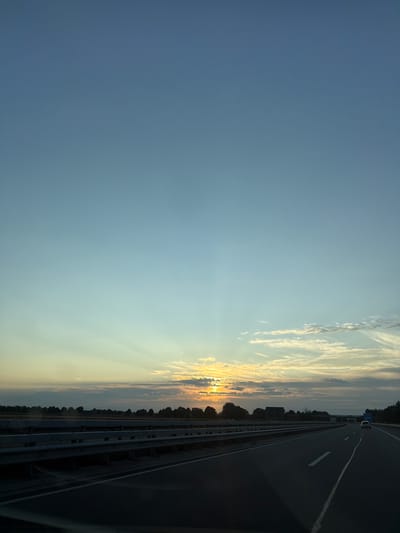 A low-angle, wide shot captures a serene sunset over a highway in Bunde, Germany. The sky is a gradient of pale blue, transitioning to soft yellow and orange hues near the horizon, where the sun is partially obscured by wispy clouds. Subtle sunbeams emanate from behind the clouds, creating a dramatic effect. The highway stretches into the distance, with two lanes visible on the right side of the frame. A silver car is driving away from the camera on the right lane. A metallic guardrail runs along the left side of the highway, separating it from a tree line that marks the edge of the landscape. The trees are dark silhouettes against the vibrant sky. The overall atmosphere is peaceful and tranquil, evoking a sense of the end of a day.