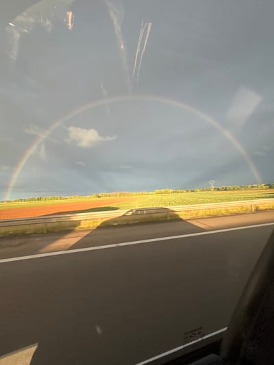 A full rainbow is visible in the sky, observed through a window. Outside the window is a road, a grassy area, and fields. The location is Kintzheim, France.