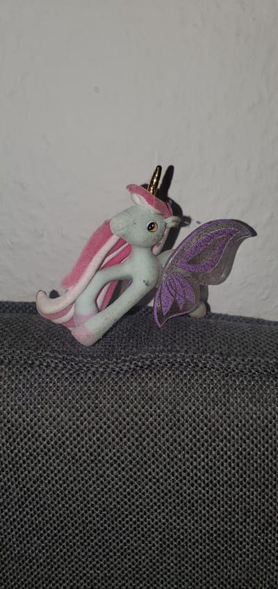 A light blue plush unicorn toy with pink and white mane and tail is positioned on a dark gray textured surface. The unicorn has a gold horn and a purple glittery wing. The background is a textured white wall. The image was taken at an unspecified indoor location.