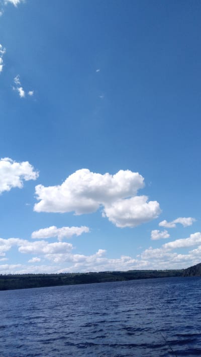 This media file depicts a serene lake scene under a bright blue sky with scattered white clouds. The foreground is dominated by the deep blue water of the lake, rippling gently, suggesting a calm day. In the middle ground, a dark, tree-covered hillside or shoreline stretches across the horizon, its dense foliage creating a strong contrast with the sky. The distant land appears lush and green.

The sky is a vast expanse of clear blue, punctuated by several fluffy white cumulus clouds. A particularly prominent cluster of clouds is near the center of the frame, appearing almost like soft sculptures. The lighting indicates it is daytime, likely late morning or early afternoon, with the sun casting bright light on the clouds and water. The overall impression is one of tranquility and natural beauty.

There are no people or identifiable objects in the frame apart from the natural elements of water, land, and sky. The scene evokes a sense of peacefulness and a connection with nature. No text is visible in the media file. The location context provided, Buzke, Ukraine, suggests a rural or semi-rural setting where such a lake and landscape would be found.