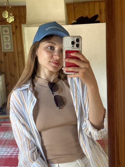 A young woman with fair skin, long brown hair, and light blue eyes takes a mirror selfie in what appears to be a room with wood-paneled walls. She is wearing a blue baseball cap with white lettering, a tan ribbed tank top, and an unbuttoned, light-colored striped shirt. Sunglasses are hanging from the neckline of her tank top. Her right hand holds a smartphone with a clear case and a red card holder attached. The background shows a glimpse of a chandelier, framed pictures on the wall, and the reflection of dark objects on a shelf. The floor is covered with a red and white plaid rug. The overall impression is that of a casual, indoor scene, possibly a home. The location in Валожынскі сельскі Савет, Belarus, is not explicitly indicated by any visual cues in the image.