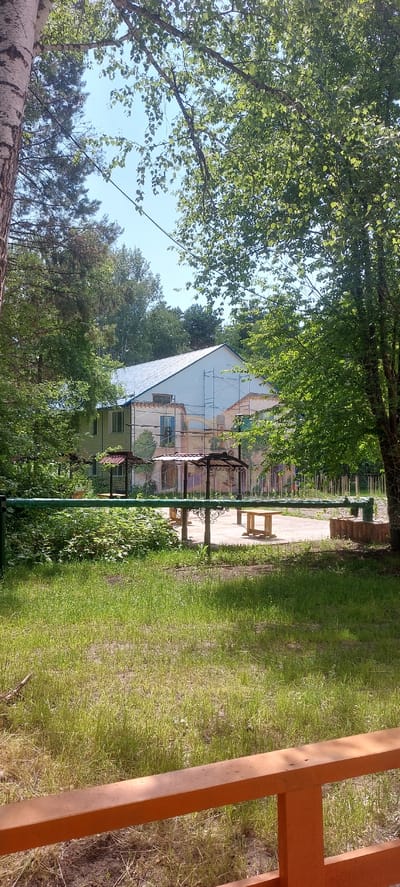The image depicts an outdoor scene likely at a recreational facility or sanatorium in Berdsk, Russia. In the background, a multi-story building with a light-colored facade and a blue roof is visible. Scaffolding surrounds a portion of the building, suggesting ongoing renovation or construction work. The building appears to have multiple windows and decorative elements, possibly murals or painted scenes, on its exterior.
In the middle ground, several covered picnic areas or gazebos with red and dark roofs are situated on a paved area. Wooden tables and benches are placed around these structures. A green metal fence runs horizontally across the foreground, separating a grassy area from the paved section.
The foreground consists of a lawn with sparse grass and some dirt patches. A wooden railing, painted orange, is prominently visible at the very bottom of the image, suggesting the viewer is standing on a porch or platform. The environment is lush and green, with numerous trees providing shade and framing the scene. The sky is a clear blue. The presence of picnic areas and a multi-story building points towards a location designed for recreation or lodging.