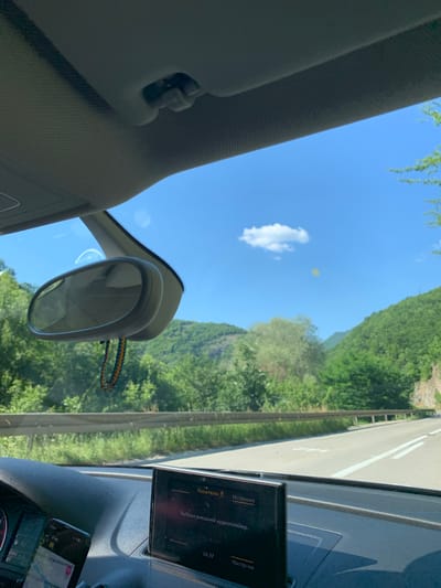 The image depicts a view from the interior of a car driving on a road in a mountainous, rural area. The sky is bright blue with a few scattered clouds. Lush green trees and vegetation cover the hillsides alongside the road. The road itself appears to be paved and has white lane markings.

Inside the car, a dashboard is visible with a central display screen showing a menu in what appears to be a Slavic language. The menu includes options like "Носители" (Carriers) and "Источник" (Source), with a selection "Выбран внешний аудиоплейер" (Selected external audio player) and "Настр-ки" (Settings). A mobile phone is mounted on the dashboard, displaying a navigation app. A rearview mirror hangs from the windshield, adorned with a colorful beaded keychain. The car's sun visor is visible at the top of the frame.

The scene suggests a journey through a scenic, natural landscape in a rural region. Given the provided location information, this is likely a road in Prijepolje Municipality, Serbia.
