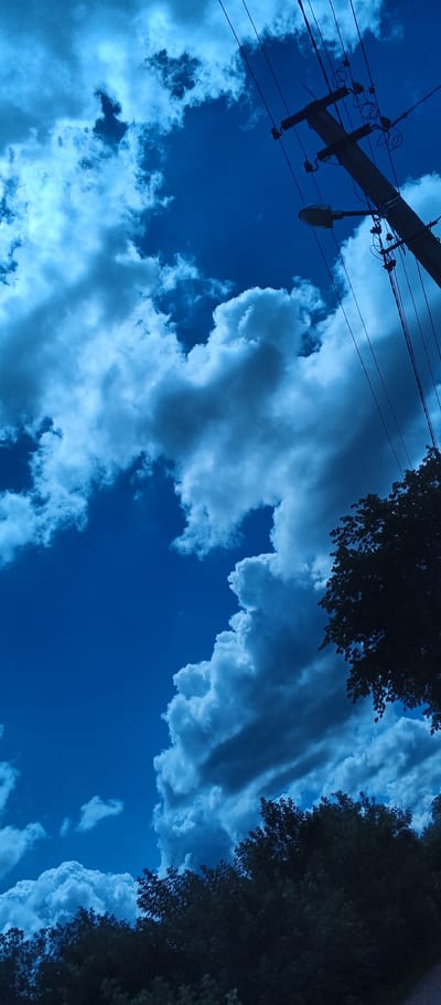 The image depicts a dramatic sky filled with large, billowing clouds against a deep blue sky. In the upper right portion of the frame, a wooden utility pole with wires and a street lamp is visible, silhouetted against the sky. The lower portion of the image shows the dark outlines of trees. The overall tone of the image is a deep blue, suggesting it was taken at twilight or during a storm. There are no people or specific actions depicted. The environment appears to be outdoors, with elements of urban infrastructure and natural foliage. The image does not contain any information or visual cues to place it specifically in the city of Popivka, Ukraine.