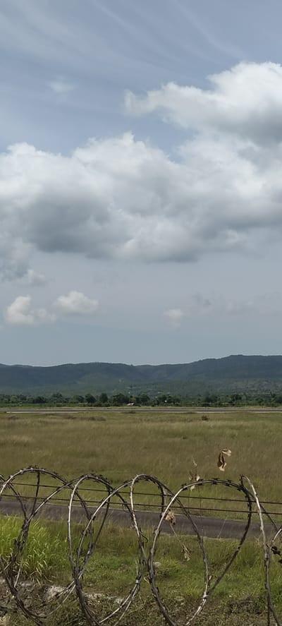 The image captures a landscape view with a barbed wire fence in the foreground, obscuring a portion of a paved surface that appears to be a runway or road. Beyond the fence, a field of dry grass stretches towards a tree line, which then gives way to a range of green, forested hills under a cloudy sky. There are no people visible in the image. The overall impression is that of a rural or undeveloped area, possibly an airport or an industrial site on the outskirts of a city. The location is specified as Buengbakjok, Indonesia.