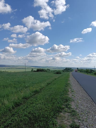The image depicts a rural roadside scene under a bright blue sky with scattered white cumulus clouds. A paved road curves to the right, bordered by a gravel shoulder and a grassy embankment on the left. In the distance, a truck with a red body and yellow load is traveling away from the viewer on the road. Beyond the road, the landscape is characterized by rolling green fields, scattered trees, and some distant structures. A power line with poles is visible running across the middle ground. The scene is consistent with a location in the vicinity of Бутурлино, Russia, given the agricultural landscape and the presence of infrastructure such as power lines and a paved road.