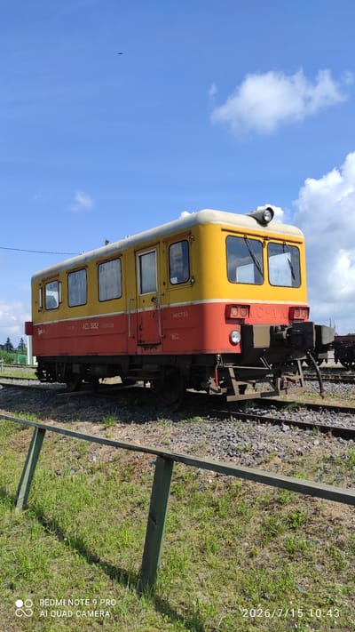 A yellow and red train car is parked on a gravel track with green grass in the foreground. The sky is blue with some white clouds. An airplane is visible in the sky. The train car has several windows and appears to be old. There is text on the side of the train car which is not legible. The image appears to be taken in a rural area of Belarus.