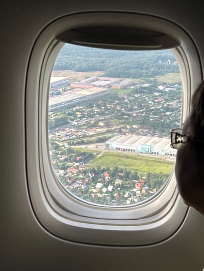 An aerial view through an airplane window captures a suburban area with a mix of industrial buildings and residential houses. The foreground shows the edge of the airplane's interior, with the curved window frame partially obscuring the scene. A person's head and shoulder, wearing glasses, are visible on the right side of the frame, looking out the window. Below the window, a long, light-blue industrial building with a white roof dominates the middle ground, surrounded by green fields and smaller buildings. Further in the distance, a dense forest forms the backdrop, with a strip of industrial development featuring long, red-roofed buildings to the left of the main industrial structure. The scene suggests a location on the outskirts of a city, likely in an area that combines industrial activity with residential development. The view corresponds to the city of Nochvino, Russia, characterized by such mixed-use landscapes.