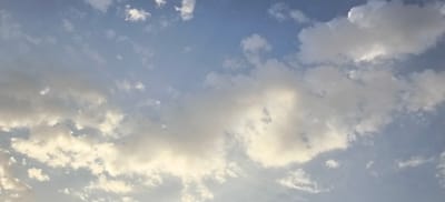 The image shows a cloudy sky above Margilan City, Uzbekistan. The clouds are a mix of white and gray, with some areas illuminated by sunlight. The sky is a pale blue. There are no people, subjects, or objects visible in the image. The scene appears to be a natural landscape.