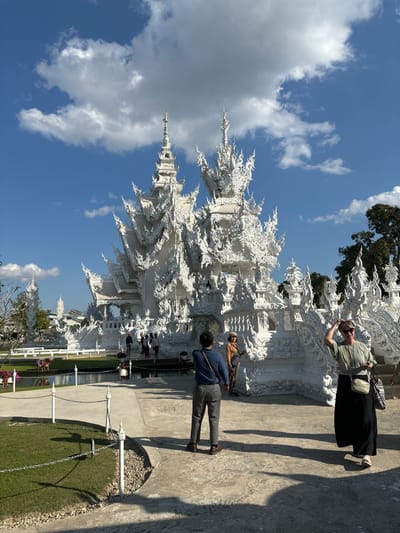 The image depicts Wat Rong Khun, also known as The White Temple, located in Ban Rong Khun, Thailand, under a clear blue sky with scattered white clouds. Bright sunlight casts distinct shadows across the scene.
The temple, a prominent, intricately designed Buddhist structure, occupies the mid-ground. It is entirely white, adorned with elaborate stucco work, tiered roofs, multiple spires, and reflective mirrored glass elements that shimmer in the sun. A pathway, bordered by decorative white posts and chains, leads towards the temple, crossing over a visible pond or moat with dark water. Several individuals are present near the temple's entrance and on its grounds, some observing or walking.
In the foreground, on a light-colored paved surface, three individuals are visible. A male subject stands with his back to the viewer on the left, wearing a blue long-sleeved shirt and grey trousers, holding a smartphone horizontally as if photographing the temple. To his right, a female subject wears a light brown jacket, patterned trousers, and a beige hat, looking towards the camera. Further to the right and slightly in front, another female subject wears a light green top, a long black skirt, and a white cross-body bag; her right hand is raised, touching her head, and she looks generally towards the left. Green grass areas are visible on the left side of the foreground, and trees are present in the background to the right.