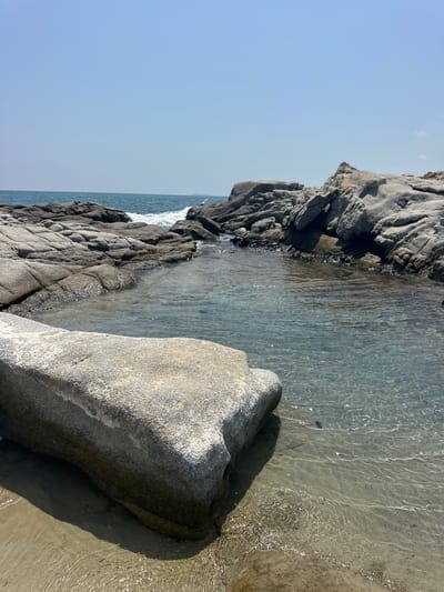 A wide-angle shot shows a rocky coastline on a clear day. The foreground is dominated by a large, rough-hewn boulder on a sandy shore. To the right and center, a calm, clear pool of water is surrounded by dark, jagged rocks. Beyond this inlet, the sea stretches to the horizon, with small waves breaking against a rocky outcrop. The sky is a pale blue, with a faint haze. The overall scene suggests a serene and natural coastal environment.