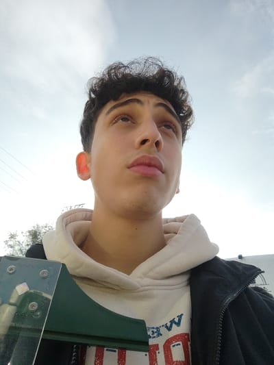 An outdoor, low-angle photograph captures a young male subject in Mankat al-Hatab, Syria, looking upwards towards a bright, partly cloudy sky. The individual has dark, curly hair, light skin, and a neutral expression. He is dressed in an off-white hooded sweatshirt featuring a graphic design with blue and red elements, worn beneath a dark, zippered jacket. His left ear is illuminated by sunlight, appearing reddish. A dark green object, possibly a piece of equipment or a structural element with visible fasteners, occupies the lower left foreground. In the background, thin utility lines are visible against the sky on the left, with faint outlines of distant foliage and a light-colored structure on the far right. No other persons or specific interactions are depicted.