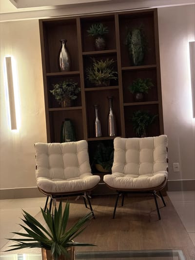 Two cushioned chairs sit on a wooden floor next to a built-in shelf filled with plants and decorative items. The chairs feature white cushions and dark metal legs. A tall potted plant stands beside one chair. Two lights illuminate the shelf and seating area. The overall ambiance is modern and cozy.