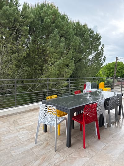 The image shows an outdoor dining area in Les Arcs, France. A long, rectangular table with a dark gray surface is surrounded by a variety of colorful chairs in yellow, red, white, and gray. The chairs are made of a patterned plastic material. A decorative metal fence separates the dining area from a large, green tree. The ground is covered in light-colored tiles. The sky is overcast.