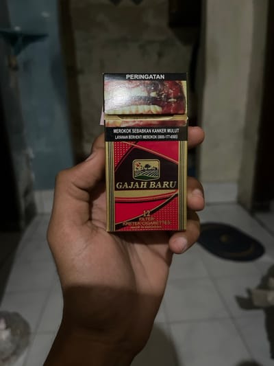 The image shows a person's hand holding a pack of "Gajah Baru" cigarettes, which are made in Indonesia. The pack is red with gold trim and features a graphic warning about the dangers of smoking. The person's hand is well-lit, suggesting the presence of a light source nearby. The background is a blur of what appears to be a tiled floor. The location, based on the provided coordinates, is in the Kecamatan Waru area of Sidoarjo, East Java, Indonesia.