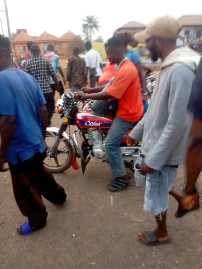 The image depicts an outdoor daytime scene on what appears to be a street or path, bustling with several people and featuring a red motorcycle prominently in the mid-ground. The lighting suggests a bright, possibly overcast day.
In the immediate foreground on the left, a person is seen from the back, wearing a blue short-sleeved t-shirt and dark, possibly brown or black, baggy pants. They are wearing bright blue slide sandals on their feet and are walking towards the right side of the frame.
Central to the image, a person is seated on a red motorcycle, facing away from the camera but with their profile partially visible. This individual is wearing an orange short-sleeved t-shirt, blue jeans, and dark slide sandals with white stripes. Their hands are on the handlebars of the motorcycle, suggesting they are either preparing to ride or are currently stationary. The motorcycle itself is red with a black seat and features chrome elements, including the front forks and engine casing. A yellow circular reflector is visible on the front fork. On the side of the motorcycle's fuel tank, the brand name "Calink" is clearly visible in white and blue lettering.
To the right of the motorcycle, another person stands, facing left towards the person on the motorcycle. This individual is wearing a light grey hooded jacket (hoodie) over a t-shirt, and light blue distressed denim shorts. They are also wearing dark slide sandals. A light brown baseball cap covers their head, and they have a visible beard.
Behind the person on the motorcycle, a partial view of another person in a blue top is visible, possibly a passenger or someone standing close by. Further into the background, several other people are walking or standing. One person in the mid-background wears a plaid shirt, another a white shirt, and others are in various darker attire.
The background features several light-colored buildings with distinct, possibly tiled, roofs, some of which have a rounded or dome-like shape. A tall palm tree is visible in the distance, suggesting a tropical or subtropical setting. The ground beneath the people and motorcycle appears to be asphalt or a hard-packed dirt surface. The overall atmosphere suggests a casual public gathering or daily street activity.