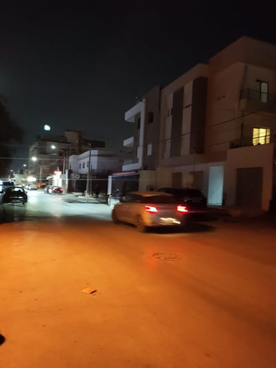 The image shows a street scene at night in an urban environment. Buildings with illuminated windows line the street, and streetlights cast a warm glow. Several cars are visible, with some in motion, indicated by blurred headlights and taillights. One car in the foreground, a silver sedan, has its brake lights illuminated. There appears to be a flag or banner with a colored light on it in the upper left portion of the image. The road surface is asphalt and appears wet, reflecting the light from the vehicles and streetlights. The overall atmosphere is that of a typical nighttime street in a residential or commercial area.