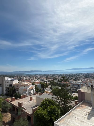 The image shows a panoramic view of the coastal city of Nea Makri Municipal Unit, Greece. The foreground is dominated by the roofs of several buildings, some of which appear to be under construction. A large evergreen tree with delicate needles stands prominently in the mid-ground. Beyond the residential area, a body of water, likely the Aegean Sea, stretches out to the horizon. Distant mountains form a hazy backdrop against a bright blue sky dotted with wispy white clouds. The overall impression is of a sunny day in a Mediterranean coastal town.