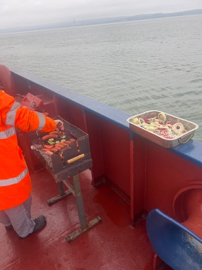 The image displays a barbecue scene on the wet, red-painted deck of a vessel, near Bangor, United Kingdom. A partially visible individual, dressed in an orange high-visibility jacket with silver reflective stripes, checkered trousers, and black clogs, is operating a portable charcoal grill. The grill contains multiple sausages, sliced zucchini, and sliced tomatoes cooking over coals. A red fuel canister rests near the person's right side. To the right, a foil-lined metal tray filled with marinated raw vegetables, including sliced onions and red bell peppers coated in a white sauce, sits on the blue-painted railing. Beyond the railing, calm grey water extends to a distant landmass featuring hills and a prominent industrial chimney stack under an overcast sky.