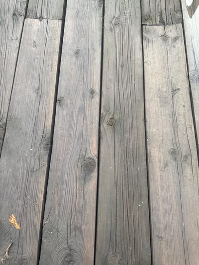 The image shows a close-up, overhead view of weathered, grey wooden planks laid side-by-side. The wood grain is prominent, with visible knots and cracks. There are scattered small debris, including what appears to be a dried leaf in the lower left corner. The lighting suggests an outdoor environment, likely during the day. The scene is devoid of any people or animals. There is no visible context to place this image specifically in Novopavlovka, Kyrgyzstan.