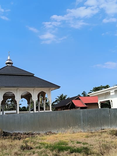 The image shows a building, likely a mosque or Islamic structure, in Aluebutuha, Indonesia. The building has a black tiled roof, white ornate columns and arches, and a silver dome with Islamic crescent symbol on top. In the background, there are two other buildings, one with a red roof and the other with a dark roof. A corrugated metal fence runs along the front of the property, and there is a field of dry grass in the foreground. The sky is blue with scattered white clouds. There are no people visible in the image.