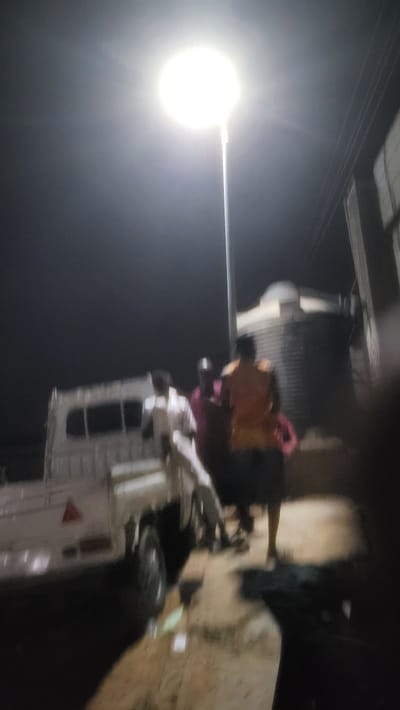 An electrical worker stands in a raised utility bucket in Jogana, Nigeria, working on a power line next to a bright street light at night. The bucket truck is positioned near a utility pole. The background is dark, and the image is slightly blurry.