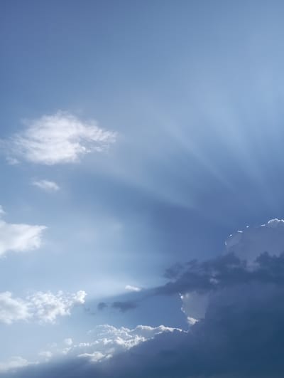 The image displays a view of the sky above Beruniy, Uzbekistan. It is a daytime scene characterized by a predominantly blue sky with scattered clouds. Rays of sunlight are visible, piercing through some of the clouds and creating a dramatic effect. The clouds vary in density, with some appearing wispy and white, while others are darker and more voluminous, suggesting potential storminess or deeper atmospheric layers. The light rays emanate from the upper right portion of the frame, casting shadows and highlighting the textures of the clouds. No people, structures, or specific landmarks of Beruniy are identifiable in the image, as the focus is solely on the atmospheric conditions.