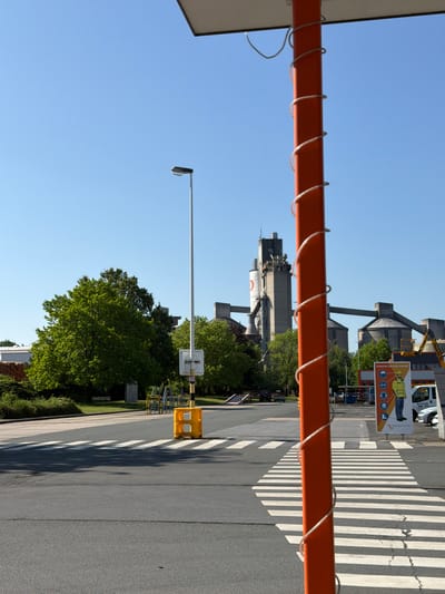 The image captures a street scene in Beckum, Germany, with a focus on an orange post wrapped in a white coil. In the foreground, a zebra crossing is visible on an asphalt road. To the left of the zebra crossing, a yellow barrier and a lamppost stand. Behind the crossing, a sign with safety instructions and an image of a construction worker is displayed. In the background, industrial buildings, including silos and cranes, dominate the skyline. Lush green trees and a clear blue sky complete the environment. The scene appears to be set during the daytime, with shadows indicating the sun's position. No people are actively engaged in any discernible actions within the frame.