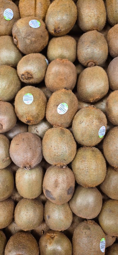 The image displays a close-up, vertical, overhead view of a large pile of kiwifruit. The fruits are oval-shaped with fuzzy brown skin. Some of the kiwifruit have round, colorful stickers on them. The lighting is even, suggesting an indoor setting, possibly a market stall or grocery store. There are no people visible in the image, and the focus is entirely on the stacked kiwifruit. The origin or specific location of this pile of kiwifruit within Navalcarnero, Spain, cannot be determined from the visual information provided.