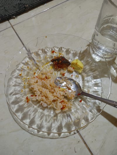 A close-up, overhead view shows the remains of a meal on a clear, patterned glass plate, resting on a tiled floor. A portion of rice, mixed with chili flakes and what appears to be a piece of fish, is visible on the plate. To the side of the rice is a dollop of yellow food, possibly egg or a condiment. A silver spoon rests on the rice, with its handle extending to the right. Next to the plate, a glass of clear liquid, likely water, is partially visible. The background consists of light-colored floor tiles and a dark, textured rug. The scene suggests the meal has been partially consumed. The location of this scene is believed to be in the city of Lhoksukon, Indonesia, based on the provided context.