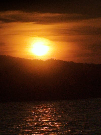 The image depicts a sunset over a body of water, likely a river or lake, with a silhouetted landmass in the background. The sky is filled with warm hues of orange and yellow, with the sun appearing as a bright, luminous orb. The water reflects the colors of the sky, creating shimmering streaks of light on its surface. The landmass, which appears to be a hillside or densely vegetated area, is dark and indistinct due to the low light conditions. The overall impression is one of a tranquil and atmospheric natural landscape. There is no indication of the city of Ustia, Moldova, in this image.