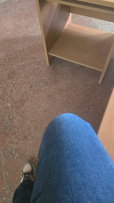 The image shows a person's legs, clad in blue denim jeans and a sneaker, resting on a speckled brown floor. To the right, a light brown wooden shelf unit is visible, with two shelves. The scene appears to be indoors, possibly in a classroom or office setting. The lighting suggests it could be daytime. No specific landmarks or recognizable features place the image definitively in Ukhta, Russia.
