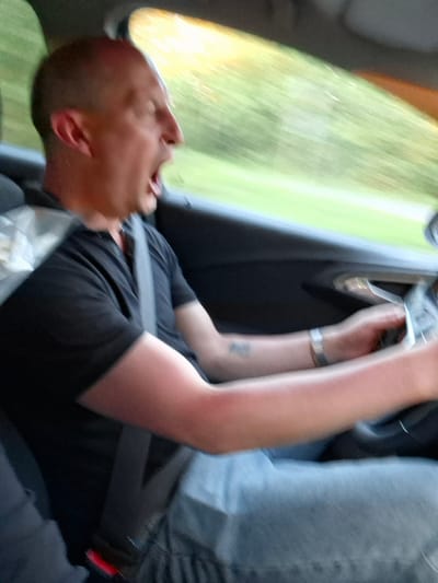 A man wearing a black shirt and blue jeans is seated in a car, secured with a seatbelt. The vehicle appears to be in motion, indicated by the blurred view of green and yellow landscape visible through the car window. The man is likely reaching for the car door handle. The image does not provide enough detail to confirm the exact location in Любанскі сельскі Савет, Belarus.