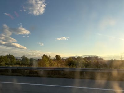 This media file depicts a scenic view captured from a moving vehicle, likely on a highway, near Mukhrani, Georgia. The scene is set during the golden hour, with the sun low in the sky, casting a warm, yellow-orange glow across the landscape. The weather appears clear with scattered clouds, which are illuminated by the setting sun.
The foreground features a road with a white lane marker and a reflective guardrail, indicating travel. Beyond the guardrail, a grassy verge leads to a dense line of trees and bushes. In the middle ground, there are more trees and vegetation, with hints of cultivated fields or open land interspersed. A few structures, possibly buildings or industrial elements, are visible in the distance, along with tall power lines and pylons, suggesting the presence of a settlement or infrastructure. The background is dominated by rolling hills or mountains, softly silhouetted against the bright sky.
The overall mood is serene and peaceful, characteristic of a late afternoon or early evening drive through the countryside. The sunlight creates lens flares and a soft, hazy effect, adding to the atmospheric quality of the image. There is no visible text in the media file.