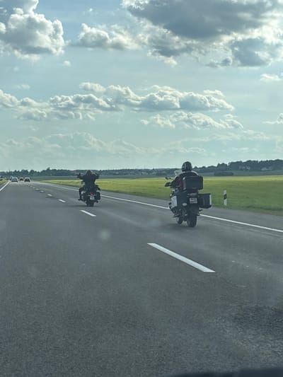 This media file, captured in Лугаваслабадскі сельскі Савет, Belarus, shows a sunny day with scattered clouds, suggesting it's daytime. The scene is a road with two motorcycles, each carrying a rider wearing a helmet. The motorcycles are equipped with luggage carriers, indicating they are likely on a journey or trip. The road is paved and marked with white lines. To the left of the road, there is a grassy field bordered by trees in the distance. The overall mood appears to be one of travel and freedom, with the open road and clear sky contributing to a sense of adventure. The license plate of the motorcycle in the foreground is partially visible, showing some letters and numbers.