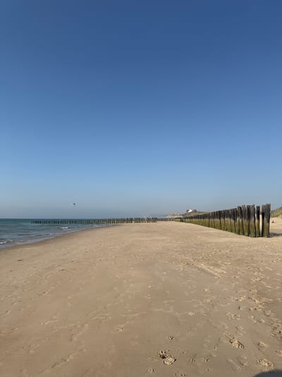 The image depicts a beach scene in Wissant, France, under a clear blue sky, suggesting fair weather and likely daytime. The perspective is from the beach, looking towards the ocean and a line of wooden breakwaters that extend into the water. The sand is light brown and shows numerous footprints, indicating recent human activity.

In the distance, a small, white kite can be seen flying over the water, suggesting a recreational activity or event taking place. The breakwaters are a prominent feature, composed of vertical wooden posts, some of which appear weathered and covered in algae or moss. Beyond the breakwaters, the ocean is visible with gentle waves lapping the shore. In the far distance, along the coastline, there are hints of buildings and dunes, providing a sense of the surrounding landscape.

The image is oriented vertically, with the sky dominating the left side of the frame and the beach and ocean occupying the right. The composition emphasizes the vastness of the sky and the texture of the sand. No text is visible in the image. The overall mood is peaceful and serene, capturing a moment of a pleasant day at the beach.