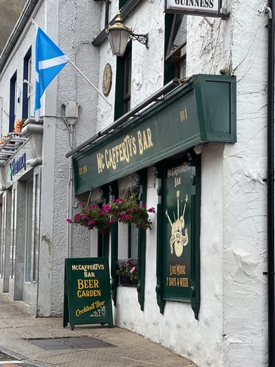 The image shows the exterior of McCafferty's Bar, a pub located in the city of Donegal, Ireland. The pub has a white exterior wall with green trim. A Scottish flag is visible flying from the building. A sign in front of the pub advertises "Beer Garden" and "Cocktail Bar". The pub's windows are decorated with a design of bagpipes and the words "CEOL AGUS CRAIC". Another sign above the door reads "MC CAFFERTY'S BAR". There is a black downpipe running down the right side of the building. The street in front of the pub is wet, suggesting it may have rained recently. There are no people visible in the image.